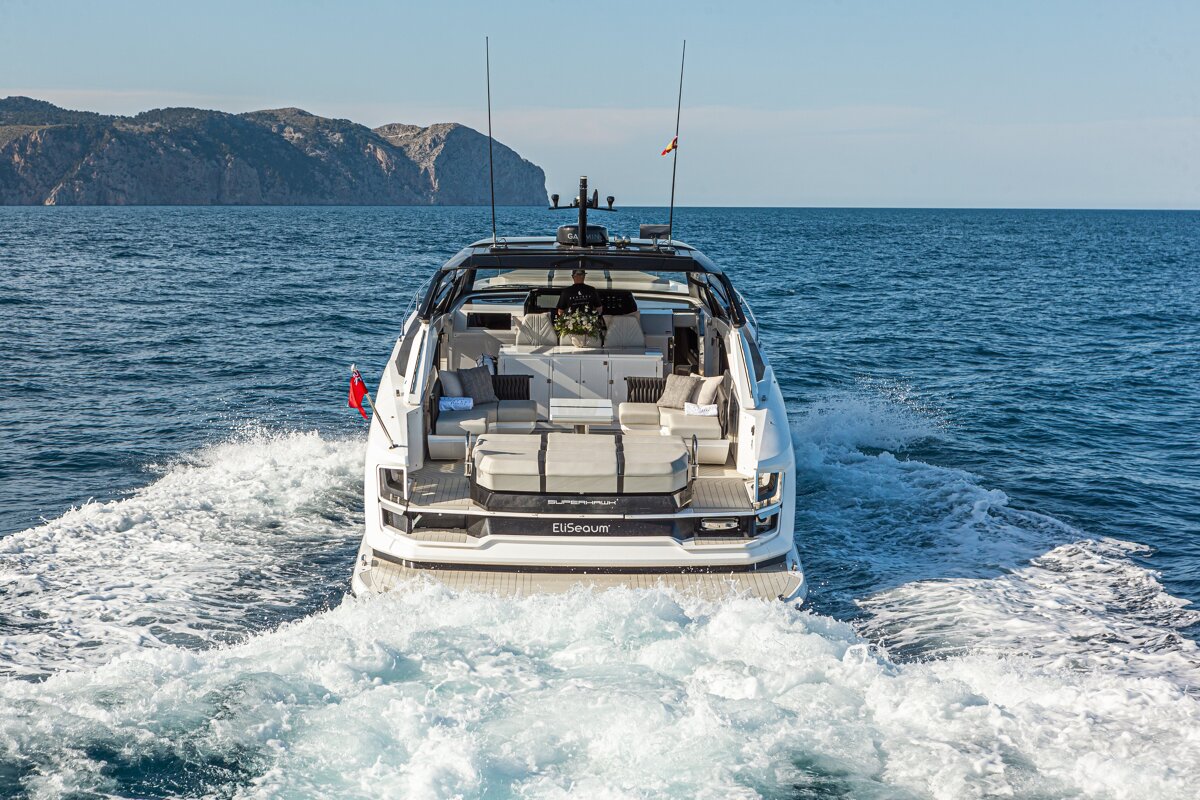 A luxurious white yacht speeds across blue water, leaving a foamy wake, with distant mountains under a clear sky on a sunny day.