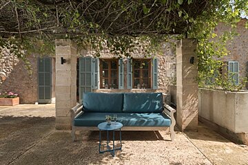 A cozy outdoor patio features a teal sofa under a leafy vine canopy, facing a stone house with blue-shuttered windows. A small table holds drinks.