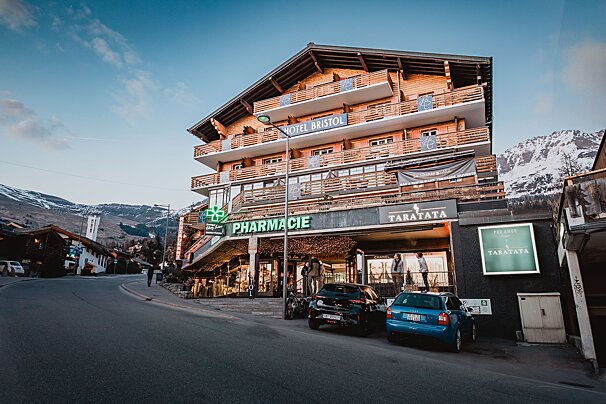 A multi-story wooden Hotel Bristol with a ground-floor pharmacy stands on a street with parked cars, backed by snowy mountains.