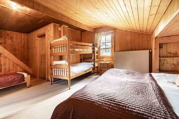 A rustic wooden bedroom with a bunk bed, a single bed, and a main bed. A window lets in light, complementing the warm wood paneling.