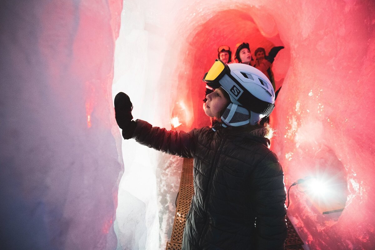 A person in ski gear explores a vibrant red and pink ice cave, touching the illuminated wall. Other people are visible further inside the unique tunnel.