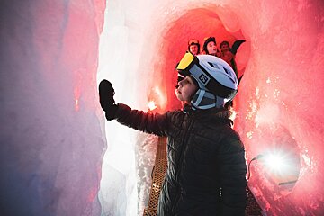 A person in ski gear explores a vibrant red and pink ice cave, touching the illuminated wall. Other people are visible further inside the unique tunnel.