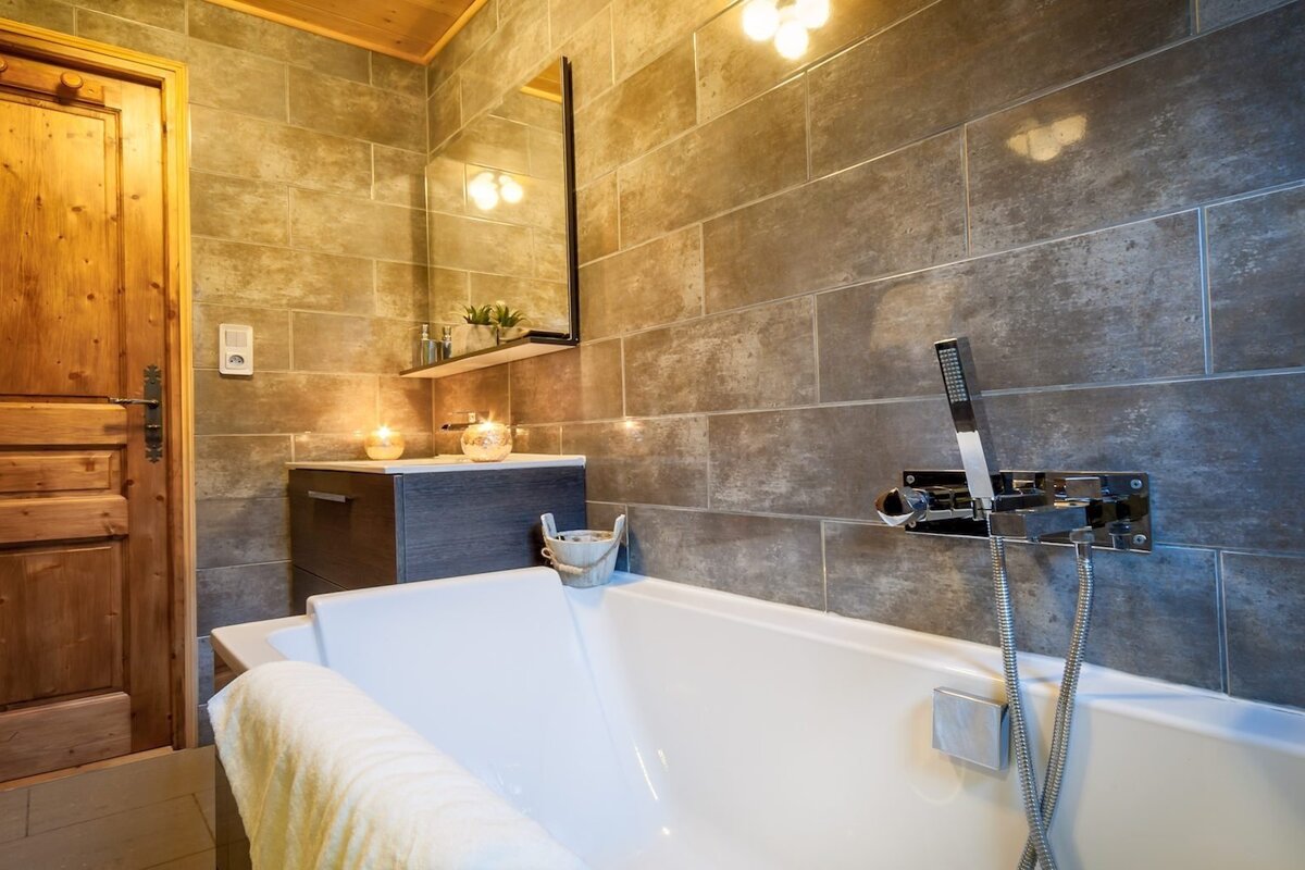 A cozy bathroom with a white bathtub, rustic tiled walls, and a wooden door. Lit candles on the vanity create a warm, inviting ambiance.