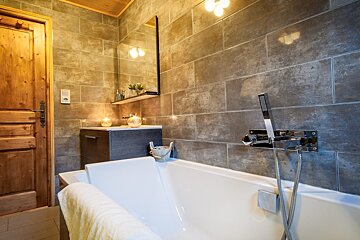 A cozy bathroom with a white bathtub, rustic tiled walls, and a wooden door. Lit candles on the vanity create a warm, inviting ambiance.