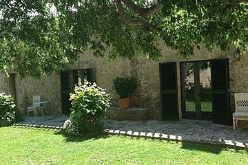 A rustic stone building with dark green shutters and doors, fronted by a lush green lawn and stone patio, all shaded by a large tree.