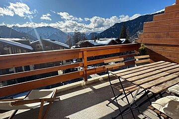 Sunny balcony overlooking a snowy mountain village and peaks under a bright, cloudy sky, with outdoor table and chairs.