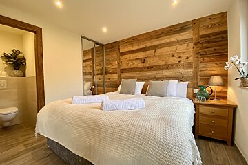 A cozy bedroom with a large bed, wood-paneled wall, mirrored wardrobe, and a toilet visible in an adjacent room. Soft lighting and plants add warmth.