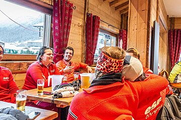 A group of ski instructors in red jackets relax with beers in a cozy, rustic wooden lodge, with snowy mountain views visible outside.