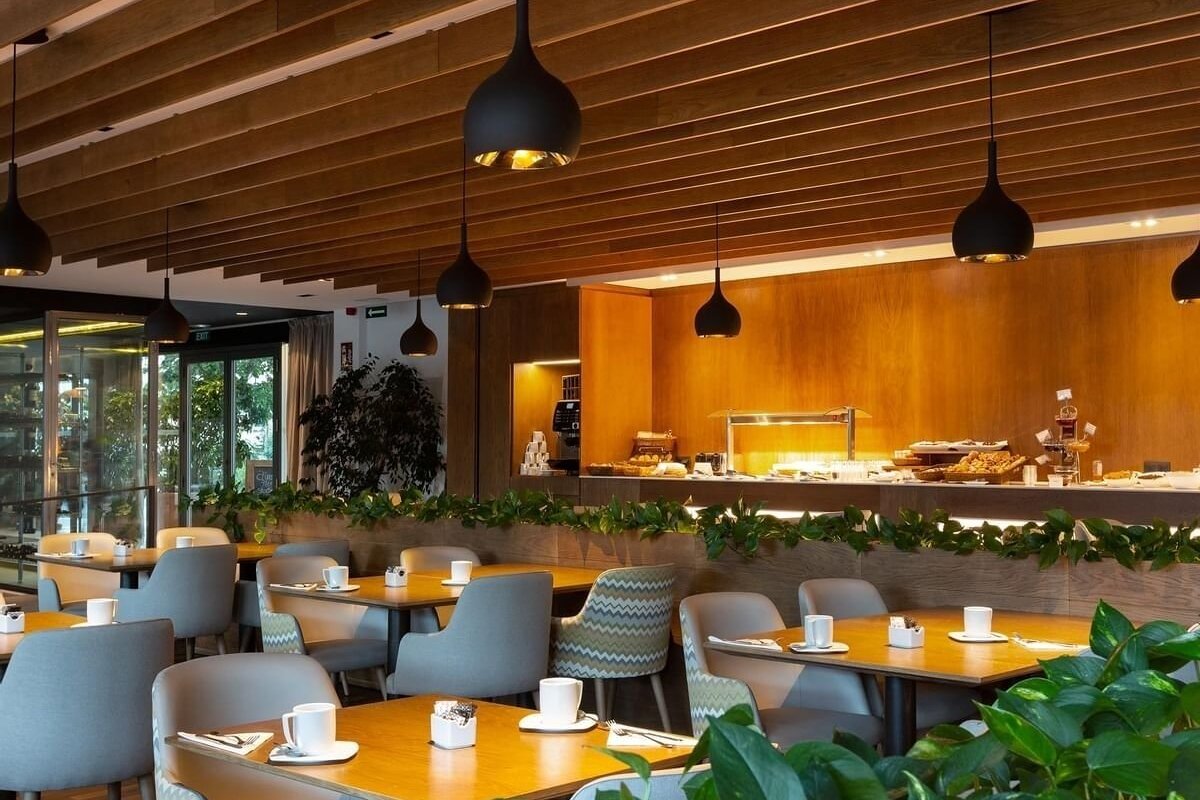 Modern restaurant interior with wood paneling, black pendant lights, set tables, a buffet counter, and green plants. Ready for dining.