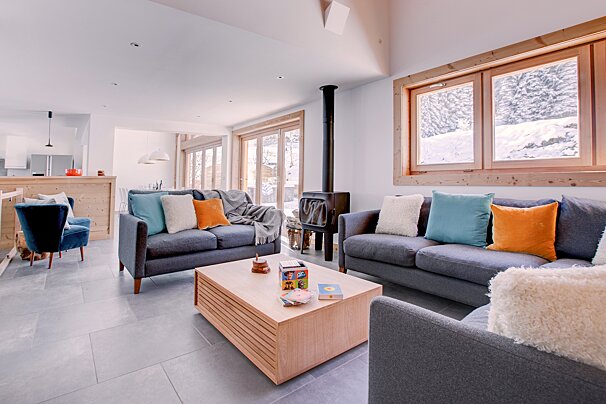 Cozy, modern living room with grey sofas, colorful pillows, a wood-burning stove, and a light wood coffee table. Snowy views outside large windows.
