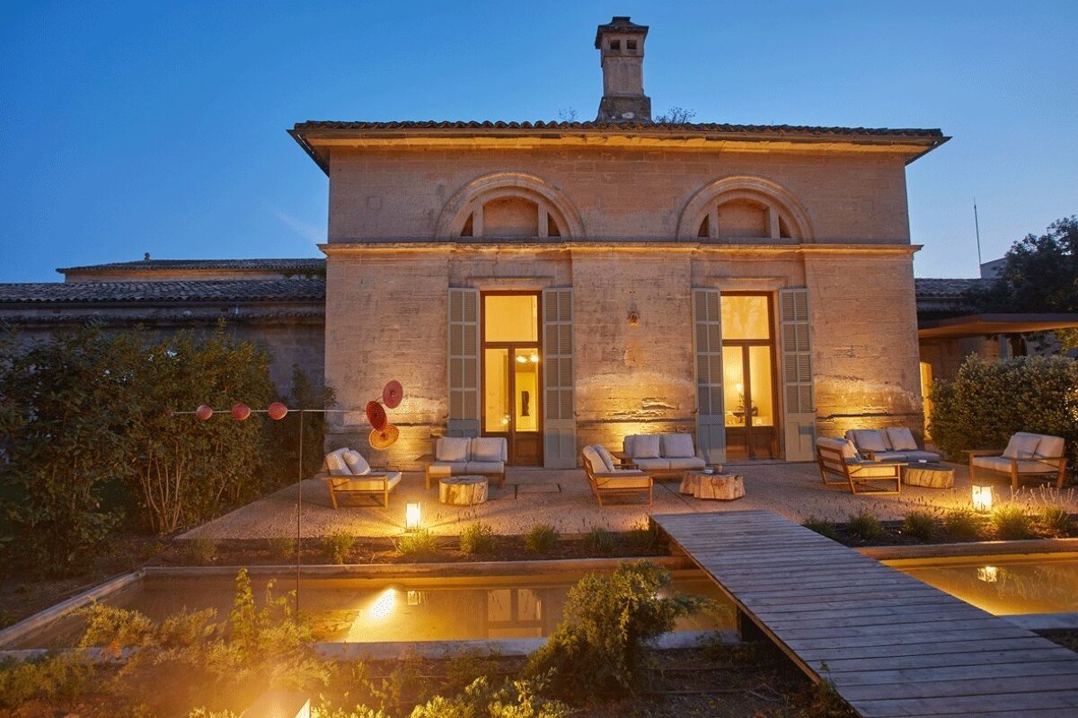 Stone house at dusk with an illuminated patio, modern outdoor seating, and a water feature with a wooden bridge.