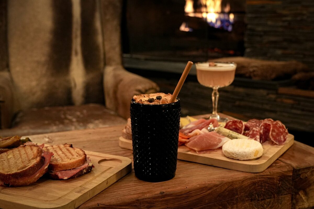 A rustic wooden table features a charcuterie board, grilled sandwiches, and two cocktails, set against a warm, cozy fireplace.