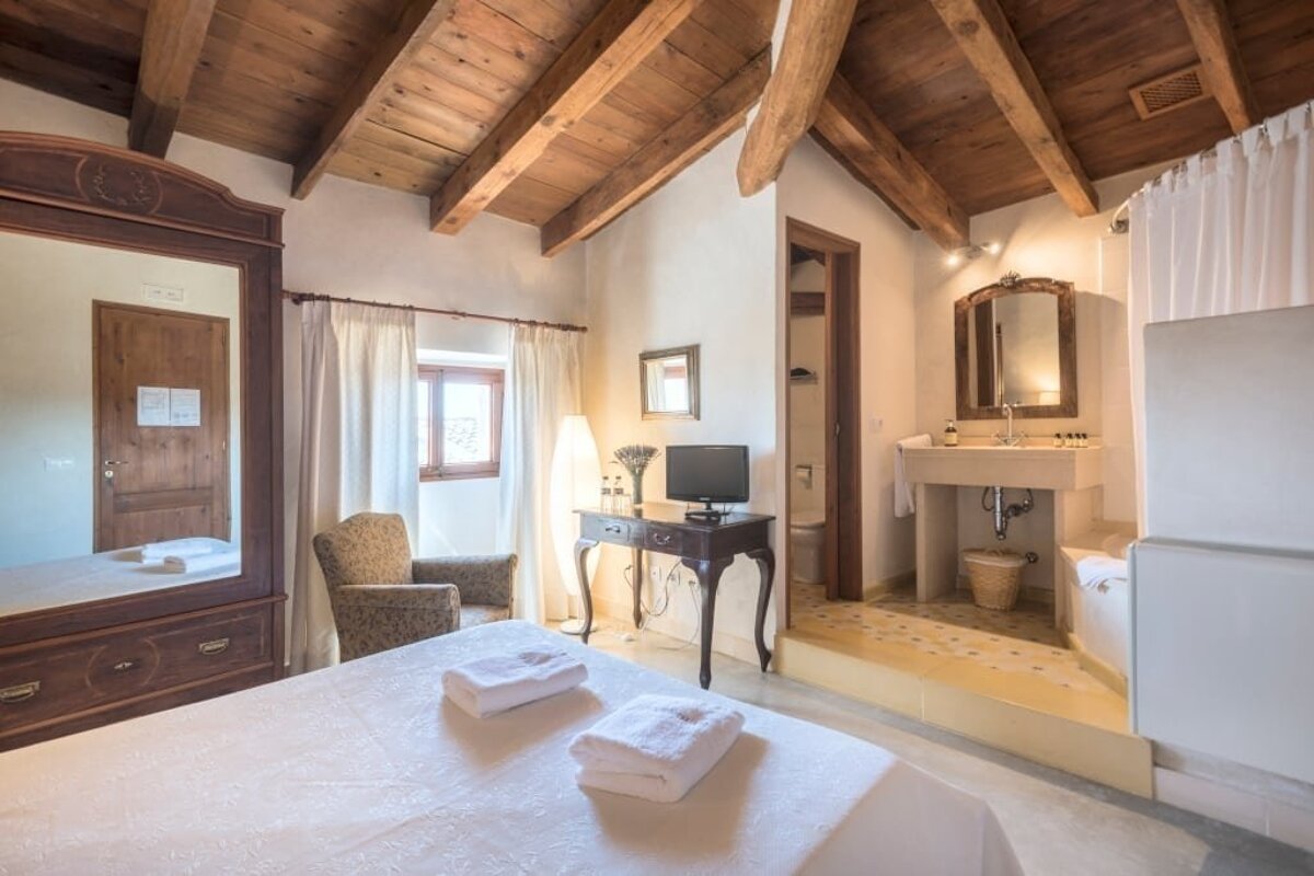 A rustic bedroom features a wooden beamed ceiling, bed, wardrobe, armchair, and an open en-suite bathroom with a sink and mirror.