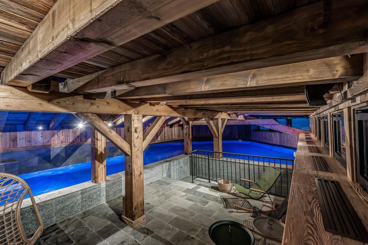 Luxurious indoor pool area with exposed wooden beams, glowing blue water, and a stone patio with lounge seating, viewed from above.
