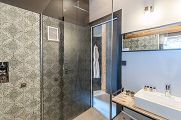 Modern bathroom with a patterned tile shower, glass partition, white rectangular sink on a wooden vanity, and a mirror above.