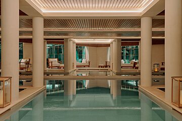 A luxurious indoor pool with elegant columns, soft lighting, and serene seating areas reflecting in the tranquil water, creating a spa-like ambiance.