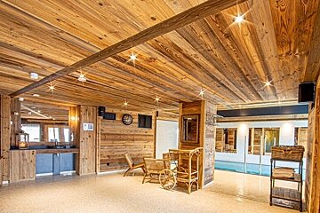 A rustic indoor pool area with wooden walls and ceiling, featuring lounge chairs, a tea cart, and an adjacent swimming pool.