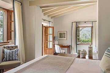 A bright, airy bedroom with white walls, exposed beams, and multiple windows offering views of lush greenery and mountains. Features a bed, desk, and rustic decor.