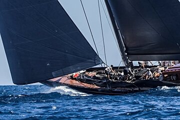 A sleek, dark-sailed yacht races dynamically through blue choppy water, its crew visible on deck under a clear sky.