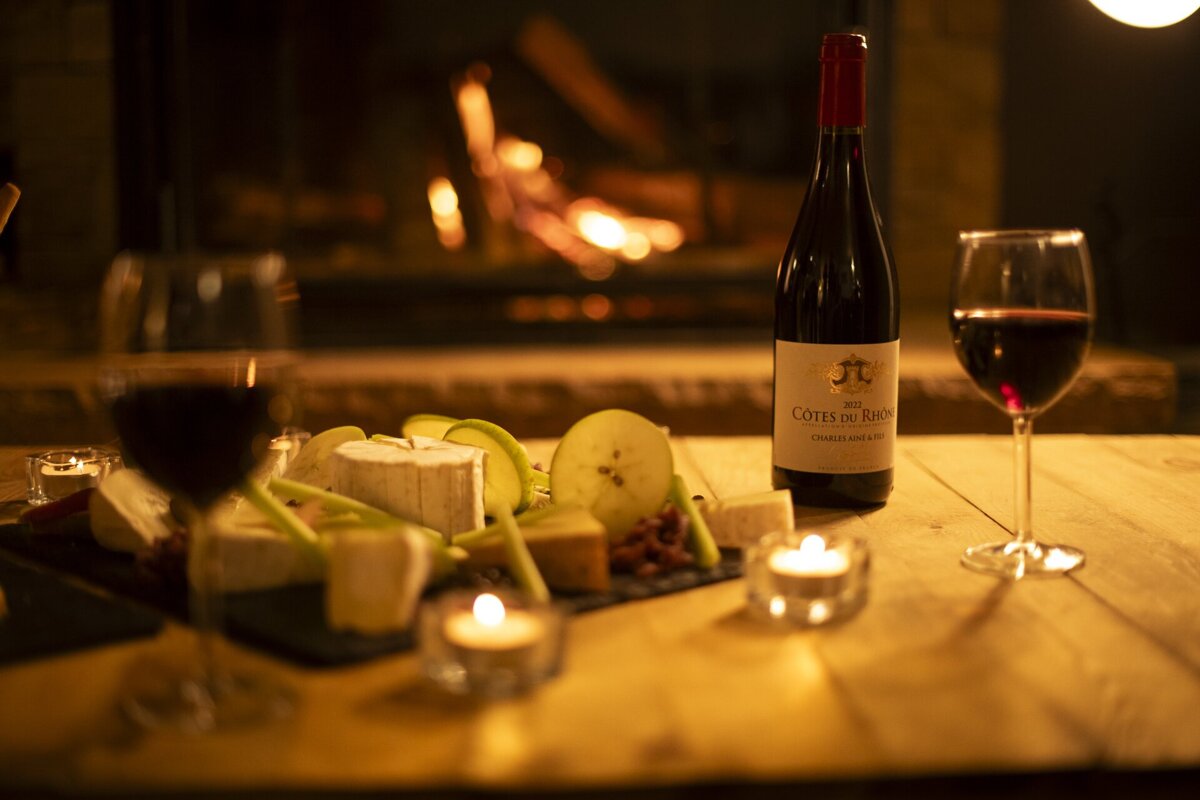 A cozy scene with a bottle and two glasses of red wine, a cheese board, and candles on a wooden table, set against a warm, blurry fireplace.