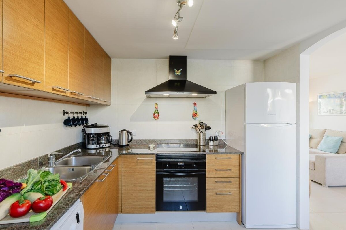 Bright, modern kitchen with light wood cabinets, granite counters, oven, fridge, coffee maker, and fresh produce. Adjoins a living area.