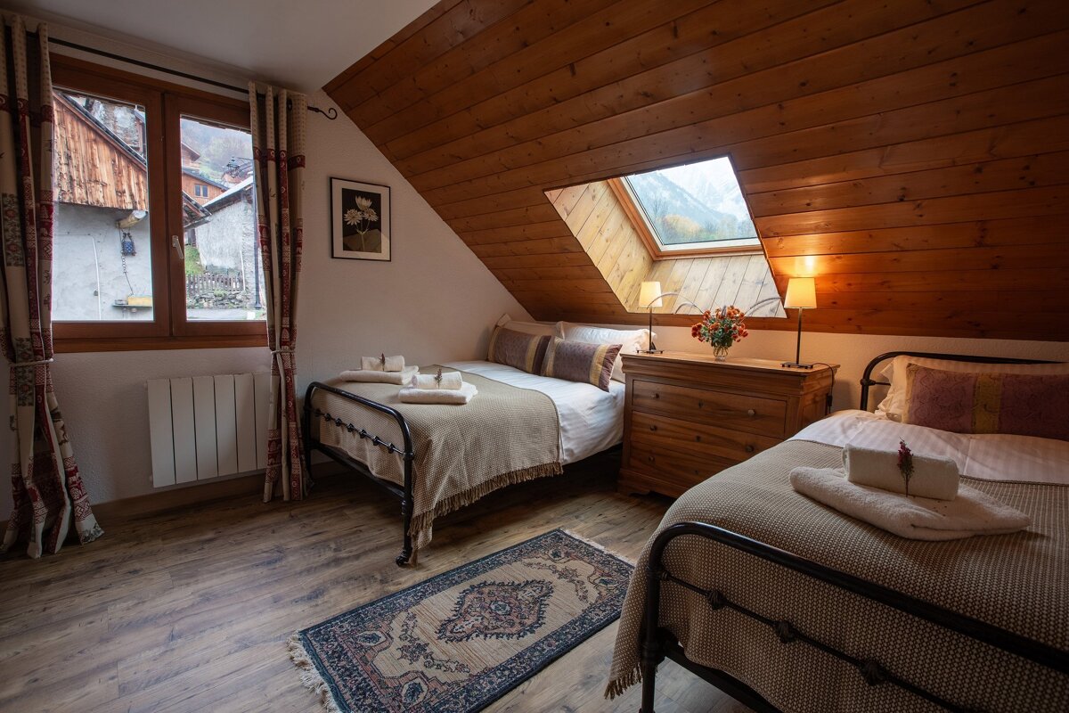 A cozy attic bedroom features two beds, a sloped wooden ceiling, and a skylight revealing mountain views. A window looks out to a village, creating a warm, inviting space.
