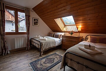 A cozy attic bedroom features two beds, a sloped wooden ceiling, and a skylight revealing mountain views. A window looks out to a village, creating a warm, inviting space.