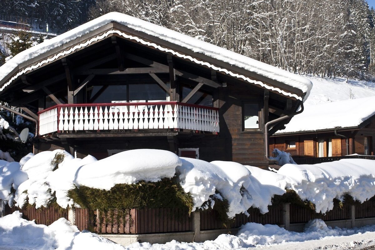 A dark wooden chalet with a balcony is heavily covered in snow, surrounded by snow-laden bushes and trees on a sunny winter day.