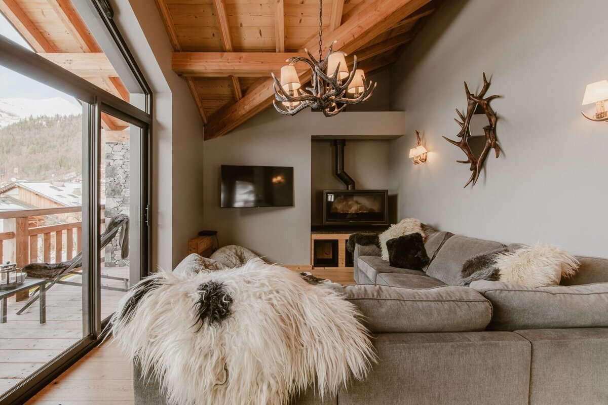 Cozy chalet living room with a wood-beamed ceiling, large gray sofa, fireplace, antler decor, and a sliding door to a mountain-view balcony.