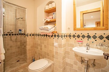 A tiled bathroom with a walk-in shower, toilet, sink with mirror, and built-in shelves holding towels and toilet paper. Decorative diamond-patterned border tiles.