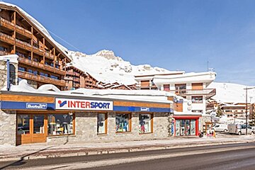 An Intersport store in a snowy ski resort, surrounded by wooden buildings and snow-covered mountains, under a clear blue sky.