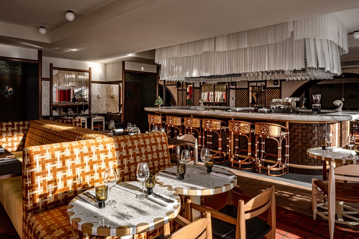 A stylish restaurant interior with warm sunlight, featuring patterned booths, marble tables, and a chic bar with rattan stools and hanging glasses.