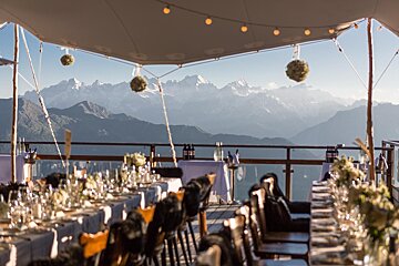 Elegant outdoor dining under a tent with string lights, offering a breathtaking view of snow-capped mountains. Tables are set.