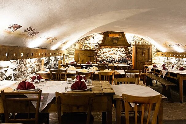 Cozy, traditional restaurant with stone walls, an arched ceiling, and wooden tables set with red napkins, illuminated by warm lighting.
