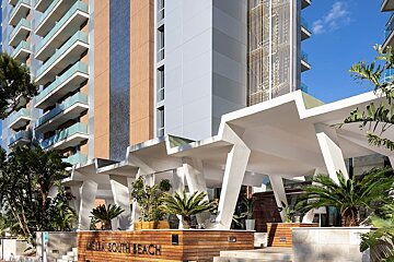 Modern hotel facade with blue-railed balconies, a distinctive white geometric canopy, lush palm trees, and a 