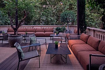 A serene outdoor lounge area with comfy brown sofas, modern chairs, and coffee tables, surrounded by lush green trees.