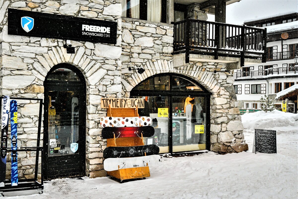 A snowy Freeride snowboard & ski shop in a stone building, displaying skis and snowboards outside on a winter day.