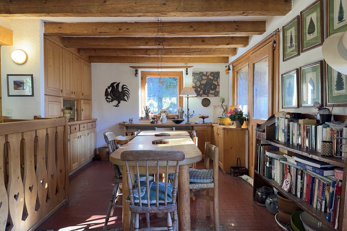 A rustic, sunlit dining area with wooden beams, light wood cabinetry, a long dining table, terracotta floor, and a bookshelf filled with books.