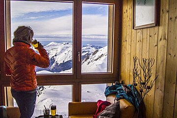 A woman in an orange jacket looks out a window at snowy mountains