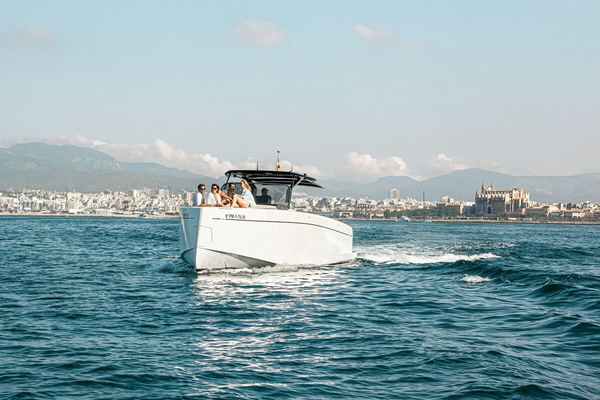 People relax on a white motorboat cruising on blue water near a coastal city and cathedral, with mountains in the background.