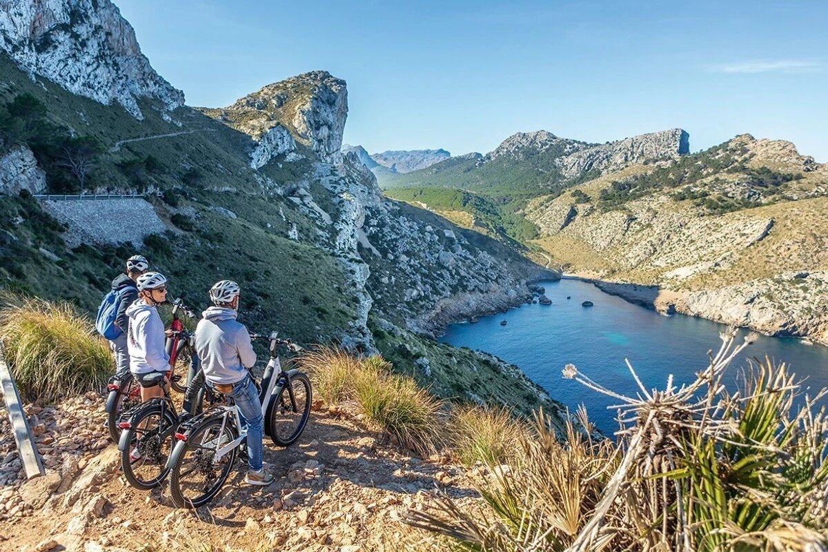 Three people on bikes admire a stunning mountain and coastal view with a clear blue cove.
