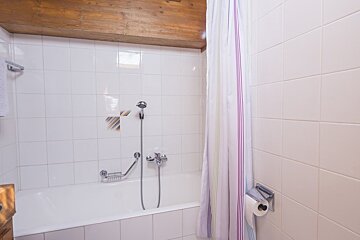 White tiled bathroom featuring a built-in bathtub with a shower attachment, striped curtain, toilet paper roll, and wooden ceiling beam.
