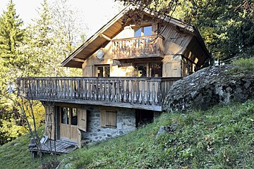 A charming wooden chalet with multiple balconies and a stone base, nestled on a grassy hillside surrounded by lush trees under bright daylight.