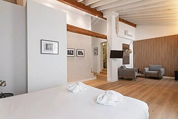 A bright, modern suite with white bedding, wood floors, exposed beams, and a seating area with gray armchairs and a wall-mounted TV.