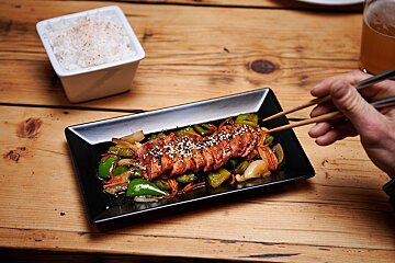 A hand uses chopsticks to eat sliced seared meat with stir-fried vegetables and sesame seeds from a black plate. Rice is nearby on a wooden table.