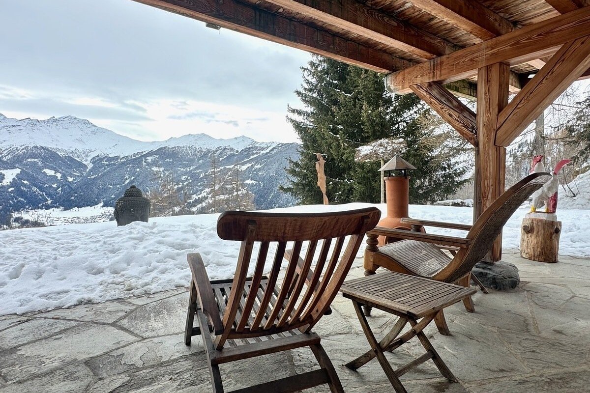 A wooden porch with chairs overlooks a snowy mountain vista and valley, with a Buddha statue nestled in the snow.