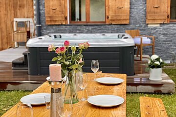 An inviting outdoor space featuring a set wooden dining table with roses and a candle, a modern hot tub, and a stone house with wooden shutters.