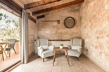 A rustic indoor-outdoor space features stone walls, wooden beams, two light armchairs, and a small table, with a view to a green balcony.