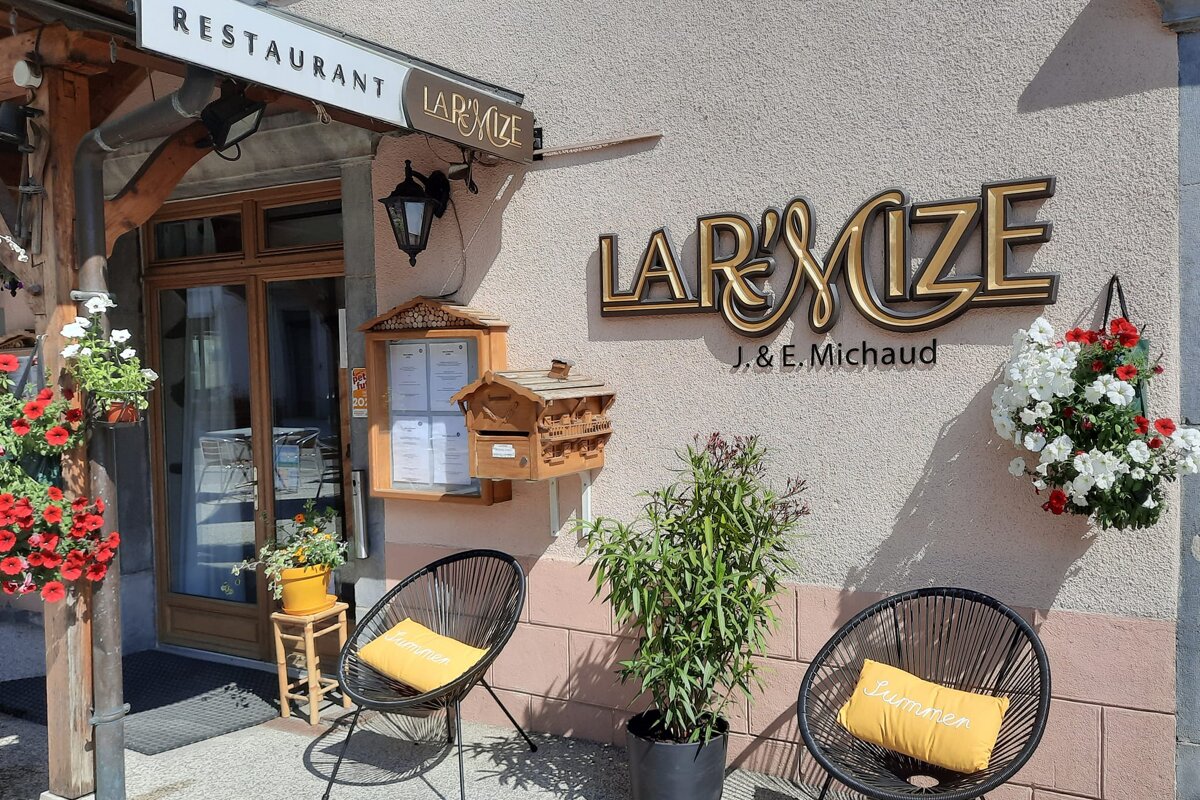 Exterior of L'Armize restaurant, featuring gold signage, hanging flowers, two black chairs with yellow 