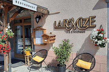Exterior of L'Armize restaurant, featuring gold signage, hanging flowers, two black chairs with yellow